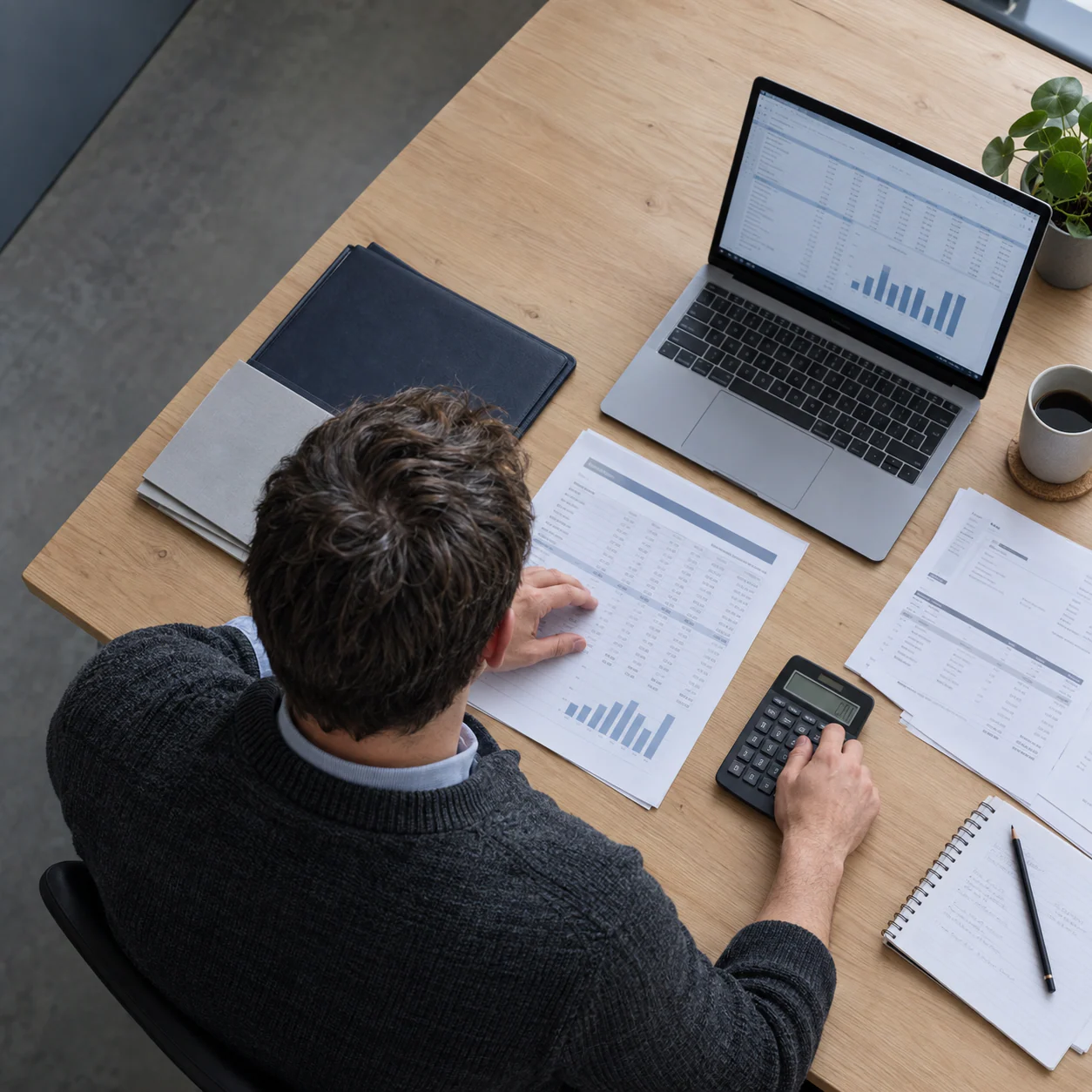 A project accountant at a desk with printed cost reports, a calculator and a laptop showing charts